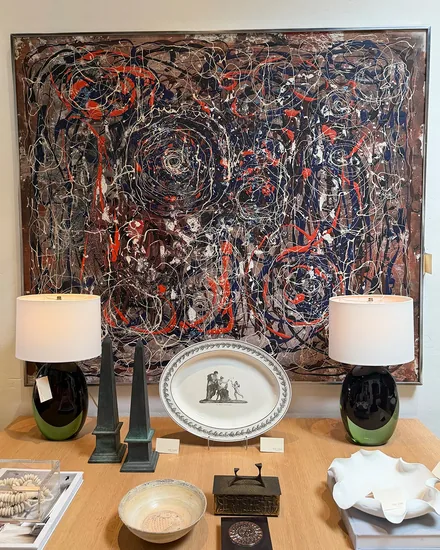 A large abstract drip painting above a console holding a pair of green and black glass lamps, bronze obelisks, a transfer printed platter and a white ruffled dish