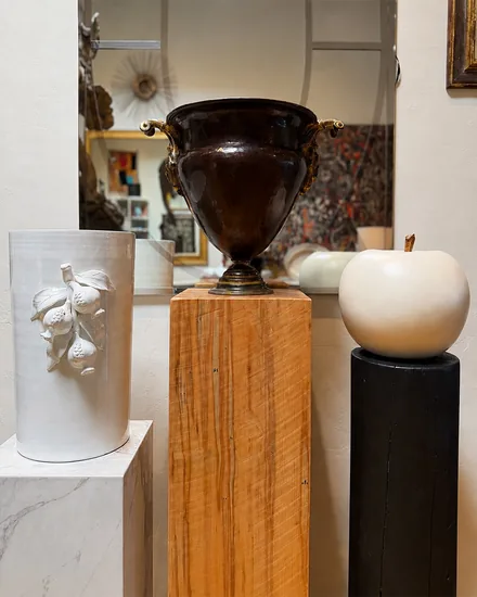 A dark glazed urn with gilt handles on a maple plinth, between a white cylinder moulded with pomegranates and a pale ceramic apple