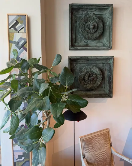 Two verdigris bronze rosette panels on the wall beside a fig tree, a narrow abstract painting and a black dome floor lamp