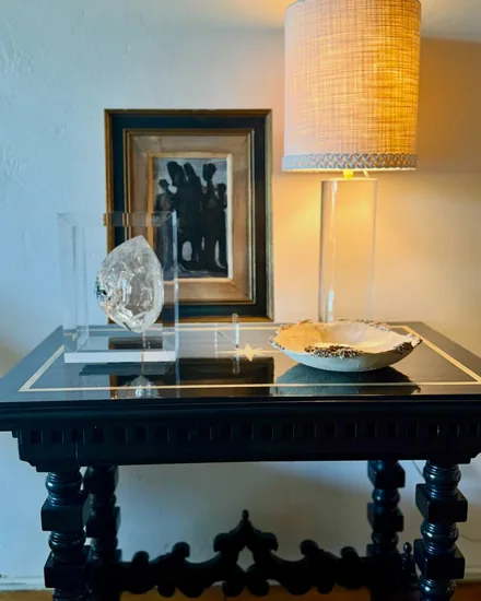 A rock crystal specimen mounted in lucite and a barnacle crusted bowl on a black turned leg table, lit by a lamp with a raffia shade