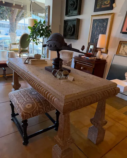 A carved travertine console by the shop window holding a carved mask on a stand and a set of silver rimmed coasters