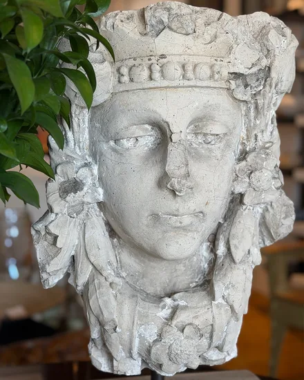 A weathered carved stone head wearing a flowered crown, mounted on a stand beside foliage