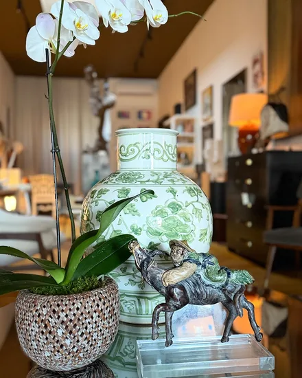 A green and white jar painted with peonies beside an orchid in a woven pot and a small painted bronze of a figure with a donkey on a lucite base